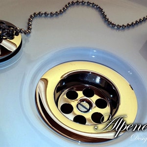 May include: A close-up of a white porcelain sink with a chrome drain stopper and chain. The drain has a circular, chrome-plated cover with multiple holes. The sink is partially visible in the background.