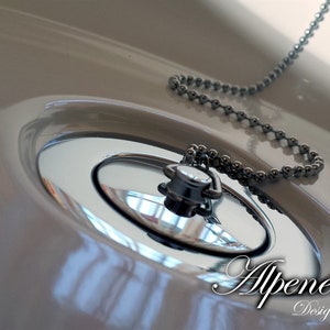 May include: Close-up of a white porcelain sink with a chrome drain stopper and chain. The drain stopper is closed and the chain is hanging over the edge of the sink. The text "Alpenemaille. Design trifft Tradition" is visible in the lower right corner of the image.