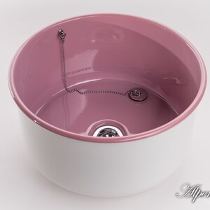 K&ouml;nnte beinhalten: Ein wei&szlig;es Keramikwaschbecken mit rosafarbenem Innenraum. Das Waschbecken hat einen Abfluss mit einer Kette und einem Stopfen.
