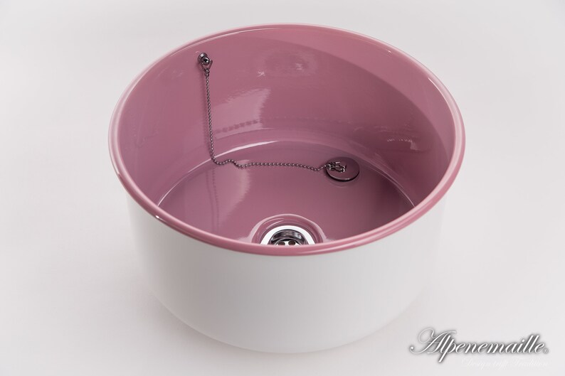 K&ouml;nnte beinhalten: Ein wei&szlig;es Keramikwaschbecken mit rosafarbenem Innenraum. Das Waschbecken hat einen Abfluss mit einer Kette und einem Stopfen.
