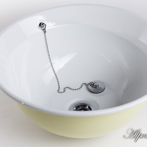 Puede incluir: Un lavabo de cer&aacute;mica blanca con un borde amarillo. El lavabo tiene un tap&oacute;n de desag&uuml;e cromado con una cadena.