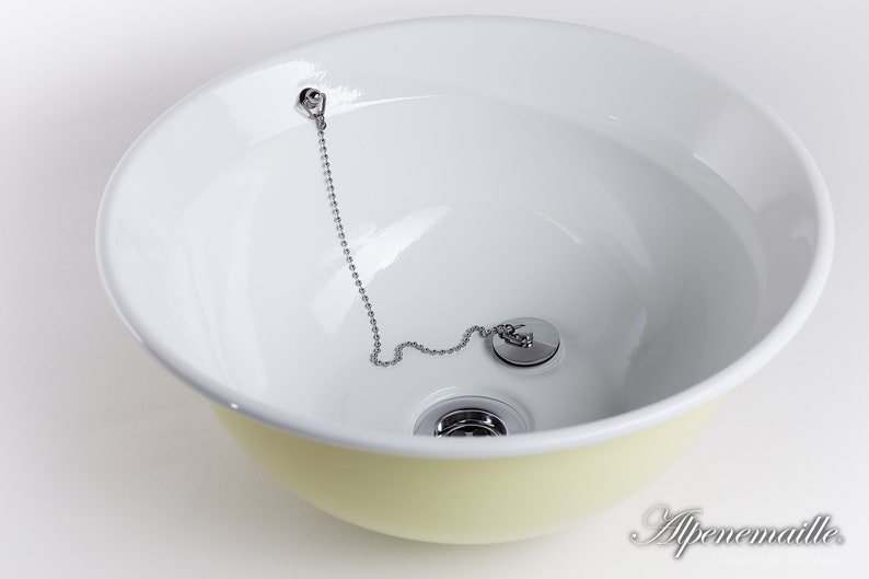 Puede incluir: Un lavabo de cer&aacute;mica blanca con un borde amarillo. El lavabo tiene un tap&oacute;n de desag&uuml;e cromado con una cadena.