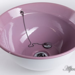 Puede incluir: Un lavabo de cer&aacute;mica rosa con un borde blanco y un tap&oacute;n de desag&uuml;e plateado con una cadena.