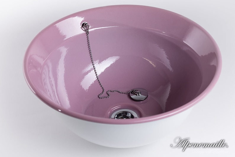 Puede incluir: Un lavabo de cer&aacute;mica rosa con un borde blanco y un tap&oacute;n de desag&uuml;e plateado con una cadena.