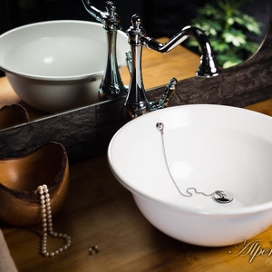 Puede incluir: Un lavabo de cer&aacute;mica blanca con un grifo cromado y una cadena unida al desag&uuml;e. El lavabo est&aacute; sobre una encimera de madera.
