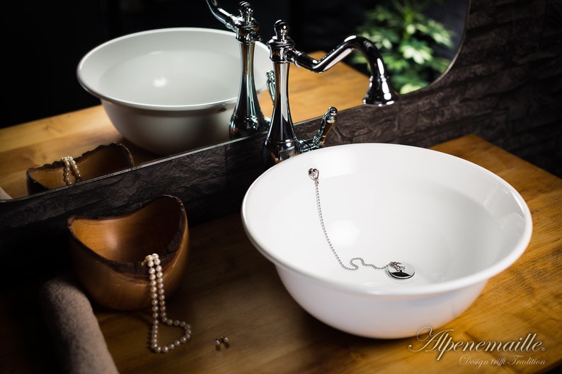 Puede incluir: Un lavabo de cer&aacute;mica blanca con un grifo cromado y una cadena unida al desag&uuml;e. El lavabo est&aacute; sobre una encimera de madera.