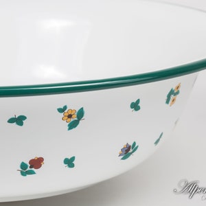 May include: White enamel bowl with a green rim and a floral pattern. The bowl has a small flower design with yellow, red, and blue accents. The bowl is made by Alpenmaille, a company that combines design and tradition.