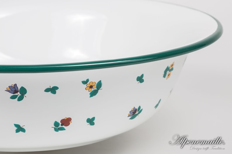 May include: White enamel bowl with a green rim and a floral pattern. The bowl has a small flower design with yellow, red, and blue accents. The bowl is made by Alpenmaille, a company that combines design and tradition.