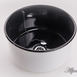 K&ouml;nnte beinhalten: Ein wei&szlig;es Keramiksp&uuml;lbecken mit schwarzem Innenraum. Das Sp&uuml;lbecken hat einen Abfluss mit einer Kette, die am Stopfen befestigt ist.