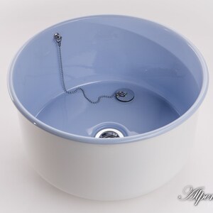 K&ouml;nnte beinhalten: Ein wei&szlig;es Keramikwaschbecken mit hellblauem Innenraum. Das Waschbecken hat einen Abfluss mit einer Kette und einem Stopfen.