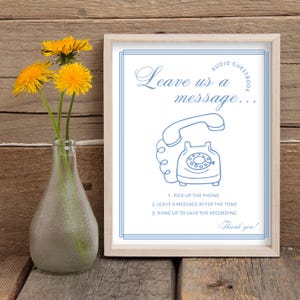 May include: A framed sign with the text "Leave us a message..." and instructions for an audio guestbook, featuring a blue illustration of a vintage telephone. A clear glass vase holds yellow flowers.