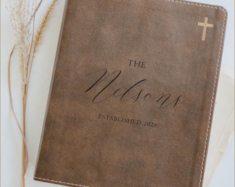 May include: A brown leather journal with the text "THE Nelsons ESTABLISHED 2026" in elegant script. A gold cross is in the upper right corner. The journal has a stitched border.