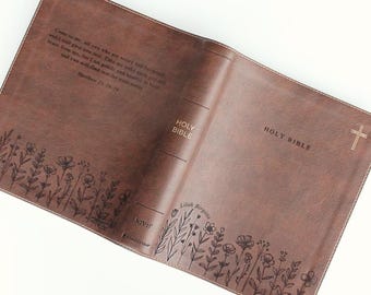 May include: A brown leather Bible cover featuring a floral line art border along the bottom edge. The front cover displays the text Holy Bible and a small cross. The back cover includes the scripture Matthew 11:28-29 and the name Lilah Birgitta.