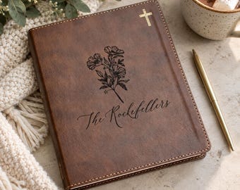 May include: A brown leather-like journal featuring a gold cross at the top, a black floral illustration in the center, and the text "The Rockefellers" engraved below. The journal has stitched edges and is accompanied by a gold metal pen.