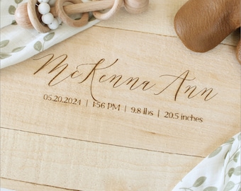 May include: A light-coloured wooden keepsake box with the name "McKenna Ann" engraved on the lid, along with birth details: 05.20.2024, 1:56 PM, 4.4 kg, and 52.1 cm. A wooden rattle and baby shoes are also visible.