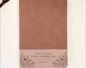 May include: A journal in a dusty rose hue, featuring a vertical ribbed texture. The lower section has a light pink band with the text "CALL TO ME & I WILL ANSWER YOU" and "jeremiah 33:3". Floral line art adorns the band. A pink ribbon bookmark is visible.