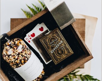 May include: A wooden box filled with playing cards, a bottle of popcorn kernels, and a small box labelled "MAST". The playing cards are a deck of "CITIZENS" playing cards with a gold and black design.