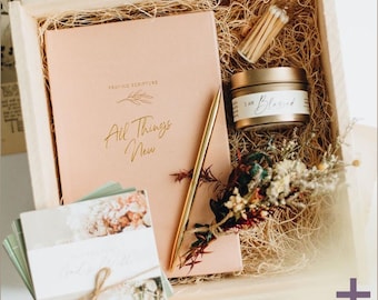May include: A wooden box filled with a pink journal with gold lettering that says "Praying Scripture All Things New", a gold pen, a small jar of candle with the text "I Am Blessed", and dried flowers.