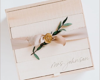 May include: A wooden box with a light brown finish, tied with a beige ribbon and a gold wax seal. A sprig of green leaves is tucked under the ribbon. The box is labelled "mrs. johnson" in a handwritten font.