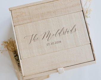 May include: A light-coloured wooden box with a sliding lid. The box is engraved with the words "The Moldstads" and the date "07.10.2026" in elegant script. The box is rectangular and has a natural wood grain finish. A white ribbon is attached to the box.