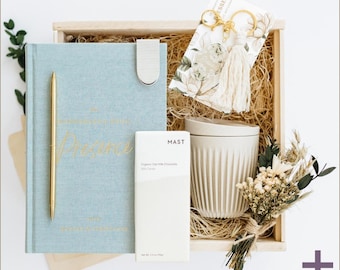 May include: A wooden gift box filled with a blue journal with a gold pen, a white chocolate bar, a white ceramic mug, a small bouquet of dried flowers, and a keychain with a tassel.