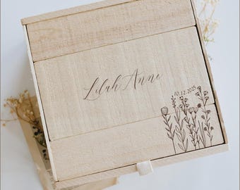 May include: A light-coloured wooden box with a sliding lid. The lid is engraved with the name "Lilah Anne", the date "10.12.2025", and a floral design. A white ribbon is attached to the box for opening.