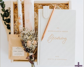 May include: A wooden gift box filled with items for self-care and relaxation. The box includes a book titled "The Wordsearch Book Becoming with Quotes & Scriptures", a rose gold pen, a small bouquet of dried flowers, a chocolate bar, and a clay face mask.