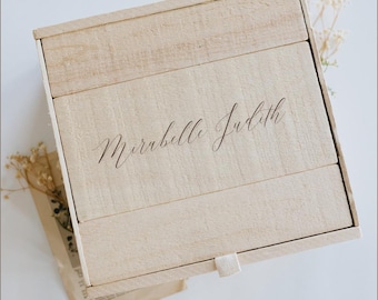 May include: A light-coloured wooden box with the name "Mirabelle Judith" elegantly inscribed on the lid. The box is rectangular and appears to be made of several wooden planks. A small, cream-coloured ribbon is attached to the front. Dried flowers and a vintage paper are in the background.
