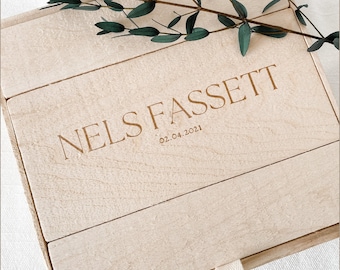 May include: A light-coloured wooden box with the name "NELS FASSETT" and the date "02.04.2021" engraved on the lid. A sprig of green leaves rests on the box. The box is made of light-coloured wood.
