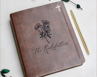 May include: Brown leather journal with gold cross and floral design. The journal has the name "The Rockefellers" printed on the cover. A gold pen rests beside the journal.
