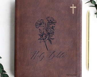 May include: A brown leather-textured Holy Bible with a dark floral line art illustration in the centre, a small gold cross in the top right corner, and the name Charlotte June printed in the bottom right corner with white stitching.