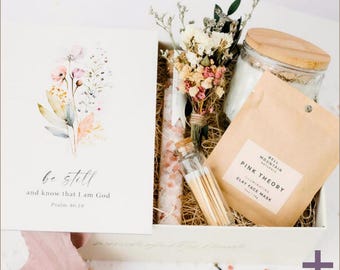 May include: A white gift box filled with self-care items, including a candle, a face mask, bath salts, matches, and dried flowers. The box also includes a card with the quote "be still and know that I am God, Psalm 46:10".