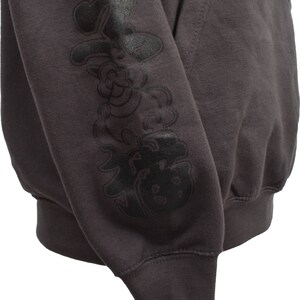 May include: A dark gray hooded sweatshirt with a black graphic print on the left sleeve. The graphic features a cartoon-like character with a large head and a small body.