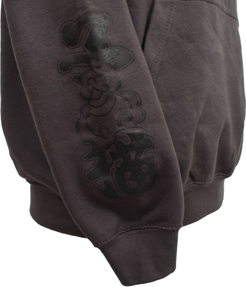 May include: A dark gray hooded sweatshirt with a black graphic print on the left sleeve. The graphic features a cartoon-like character with a large head and a small body.