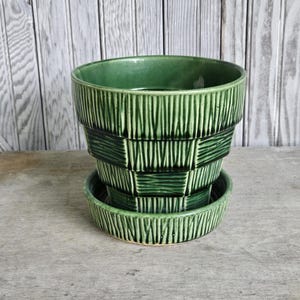 May include: A forest green ceramic plant pot with a matching saucer, featuring a textured exterior with vertical ridges and a woven basket-like pattern. The glazed finish has varying shades of green, highlighting the deep grooves in the design.