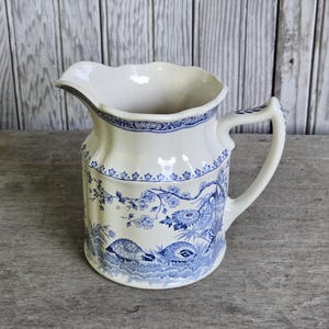 May include: A cream-colored ceramic pitcher featuring a blue transferware pattern of birds, foliage, and floral motifs. The vessel has a scalloped rim, a curved handle, and a decorative border around the upper section with a glossy finish.