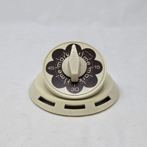 May include: A beige kitchen timer with a circular face featuring a black flower-shaped center. The dial displays the text "mark time" repeatedly around the perimeter, with numerical markings for 15, 30, and 45 minutes. A central beige knob sits atop.