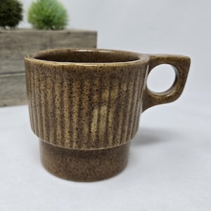 May include: Brown ceramic mug with a textured, ribbed design. The mug has a handle and a stacked base. The mug is brown with darker speckles. The mug is sitting on a white surface.