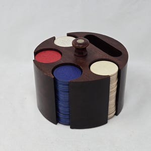 May include: A round, dark wood poker chip holder with a central handle and slots for chips. The holder contains stacks of red, blue, and off-white poker chips. The design is circular with a dark finish.