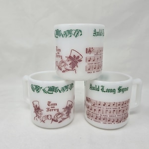 May include: Three white ceramic mugs with handles. Each mug features a different design in green and maroon. The designs include musical notes, the text "Auld Lang Syne", and illustrations of characters from "Tom and Jerry".