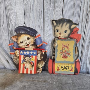 May include: Two vintage cat-shaped calendar cutouts. One cat wears a navy hat and holds a patriotic sign. The other cat has a red bow and a calendar page that reads "1941". Both are made of cardboard and have a distressed appearance.