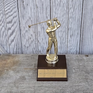 May include: A gold-colored golf trophy featuring a golfer in mid-swing, mounted on a dark brown wooden base. The base has a gold plaque that reads "WURL Mid-Winter Golf Classic 1967". The trophy is set against a weathered wood background.