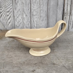 May include: Cream-colored gravy boat with a scalloped rim and a handle. The boat has a red stripe near the rim and another near the base. It sits on a round, pedestal base. The background is a weathered wooden surface.
