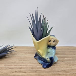 May include: A vintage ceramic planter in the shape of a seated elf, with a yellow and blue color scheme. The planter holds tall, spiky, blue-green faux plants. The planter sits on a wooden surface.