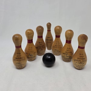 May include: Seven miniature wooden bowling pins with smooth, light brown finishes and red neck stripes, arranged in a formation around a single small, solid black bowling ball. Each pin features handwritten black ink text detailing league years.