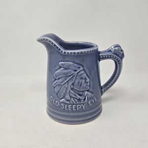 May include: A blue ceramic creamer with a raised image of a Native American in a headdress. The creamer has the words "Old Sleepy Eye" on the front.