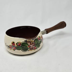May include: Cream-colored saucepan with a dark brown interior and a wooden handle. The exterior features a floral design with red, green, and brown accents. The handle is attached with a silver-colored metal piece.