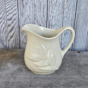 May include: Cream-colored ceramic pitcher with a curved handle and a decorative leaf design. The pitcher has a wide opening and a slightly textured surface. The background is a weathered wooden surface.