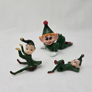 May include: Three vintage elf figurines in various poses. Each elf is dressed in a green outfit with red accents on the shoes and a matching green hat. The elves have fair skin and are in a playful, whimsical style.