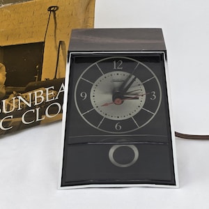 May include: A Sunbeam electric clock with a dark wood-grain top, a silver circular face featuring black numerals, and a black lower panel with a circular pendulum cutout. The clock is framed in chrome and includes a brown power cord.